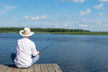 a man in a hat sits on a pier with a fishing rod in his hands and looks at the river