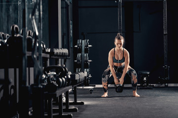 Fototapeta na wymiar Caucasian muscular brunette with earphones in ears doing workouts with kettlebell in squat position. Night workout in gym concept.