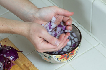 Close up of two hands gently cupping diced red onion and dropping into a bowl of water on a countertop with cutting board visible in the lower left corner.
