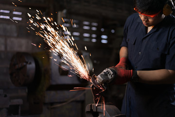 Asian male workshop engineer operating an angle grinder on a metal part with sparks flying -...
