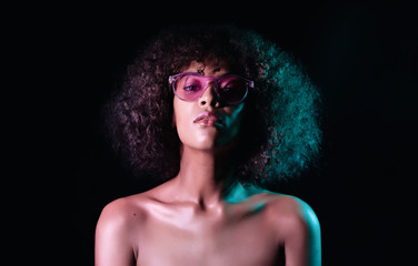 Portrait of young sexy african girl with glasses in neon light at black background. Woman with perfect makeup looking at camera and smiling. Glamour, fashion concept.