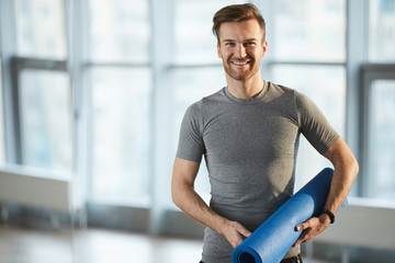 Smiling handsome young male yoga coach with cool hairstyle and stubble holding yoga mat and looking...
