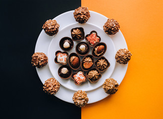 Beautiful chocolates of different shapes and fillings lie in a white plate against the background of colored paper. Orange and black color. Copy space.