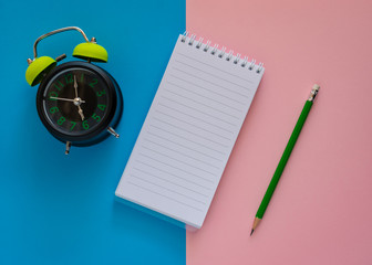 Open notebook with green pencil and alarm clock isolated on pink and blue paper background. - 260050488