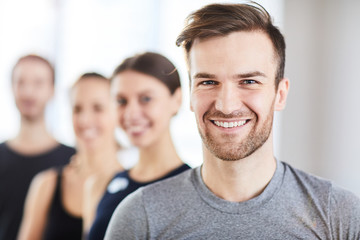 Obraz premium Portrait of cheerful handsome young guy with stubble and fashionable hairstyle smiling at camera while standing in line of fitness trainers