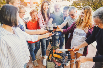 Happy family cheering and toasting with red wine glass at barbecue party - People with different ages having fun drinking and grilling meat at bbq dinner - Friendship, food and weekend activities - Powered by Adobe