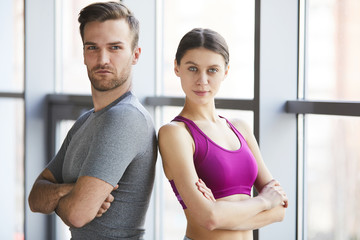 Portrait of serious confident professional fitness coaches in comfortable clothing standing back to back and crossing arms on chests