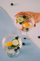 Woman holding a gin and tonic drink with blueberries, cucumber and rosemary on a blue geometric,...