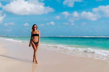 Fototapeta na wymiar Sexy tanned girl in black swimsuit and sunglasses goes on tropical beach