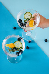 Woman holding a gin and tonic drink with blueberries, cucumber and rosemary on a blue geometric, colorful background.