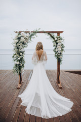 girl in a white wedding dress stands near the ceremonial arch on the background of the sea