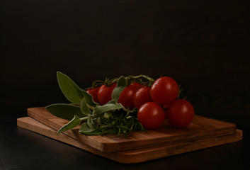 A bunch of greens for cooking meat consisting of rosemary and sage on a wooden cutting board. Nearby is a branch of fresh ripe cherry tomatoes. Tomatoes drop water. Set for cooking sauce or salad.
