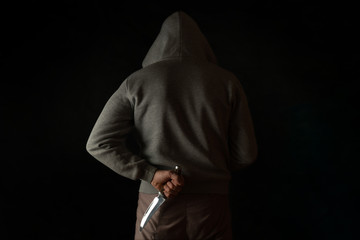 A man holds a knife in his hand behind his back, in a hood on a black background. Criminal with a...