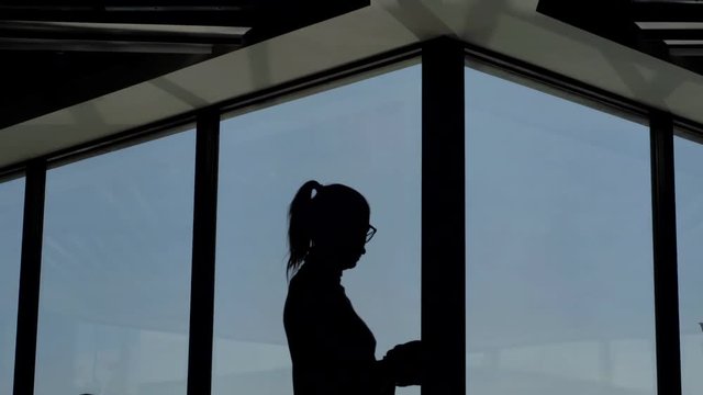 dark silhouette of adult woman standing against large window with blue sky view