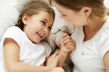 Above view portrait of happy little girl laying in bed with mom and giggling playfully, copy space