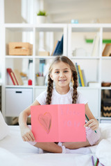 Portrait of cute little girl sitting on bed holding handmade greeting card and smiling at camera, copy space
