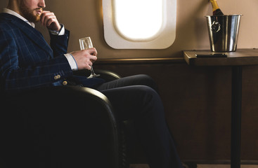 A young successful businessman in an expensive suit sits in the chair of a private jet with a glass...