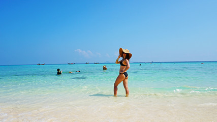 The girl in the hat and swimsuit on the beach. Glasses on. European woman on the beach with white sand. The water is turquoise with a blue tint. Photo shoot model. White sand and blue sky. Paradise.