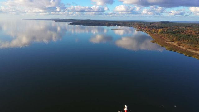 Drone shot of a small light house near Pervalka, Lithuania, surrounded by autumnal forest, near the coastline, in the calm sea with reflecting surface, on a bright sunny day