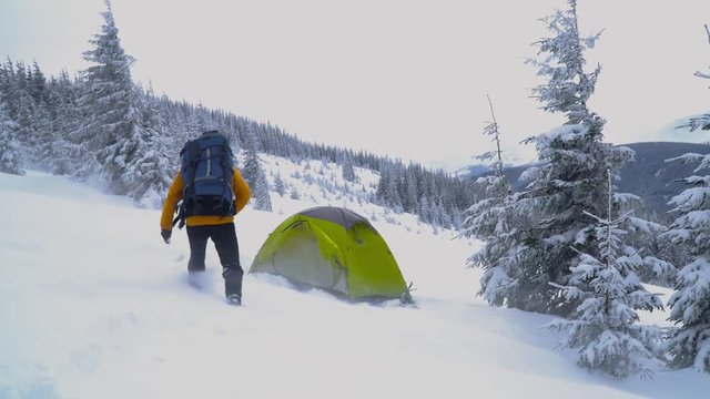 A man with a backpack travels in the winter mountains. He sleeps in a tent set up in the snow. The weather is a strong wind with snow. Carpathians. 4K
