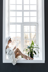 A cute young woman dressed in a white robe and with towels on her head sitting on the window sill...