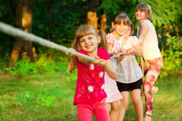 Group of happy children playing tug of war outside on grass. Kids pulling rope at park.