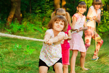 Fototapeta premium Group of happy children playing tug of war outside on grass. Kids pulling rope at park.