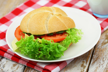 White bread roll with cheese, lettuce, tomato and cucumber on plate on checkered cloth