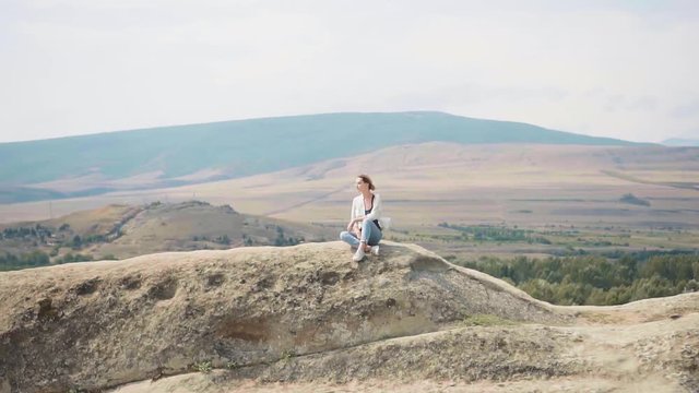 charming young woman with blond hair sits alone on top of mountain in Georgia in loose clothing, morning meditation in clear air, gorgeous nature and human as one, hills and plains alternate