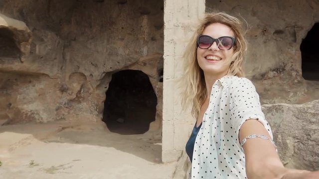 sweet young woman with blond hair flying in summer clothes and dark sunglasses, leads her husband in style follow me, wife and cameraman go to small cave in mountains of Georgia, tourists newlyweds