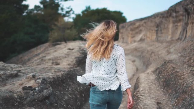 Happy woman back rear view blond hair  fly in wind walks along path in stone mountains. Girl Tourist in old town of Georgia Uplistsikhe. Person jn vacation shooting no face. casual clothing 