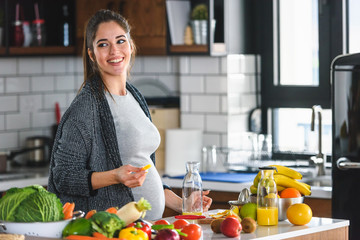 Beautiful smiling young pregnant woman preparing healthy food with lots of fruit and vegetables at...