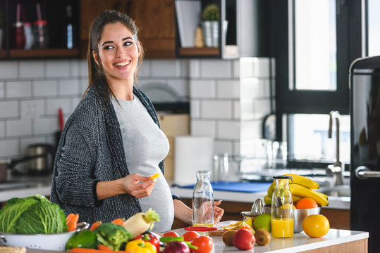Beautiful smiling young pregnant woman preparing healthy food with lots of fruit and vegetables at home kitchen