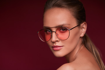 Portrait of beautiful young woman with perfect makeup wearing pink sunglasses. Smiling fashion model in aviator sunglasses posing on burgundy background. Studio shot. Summer vacation.