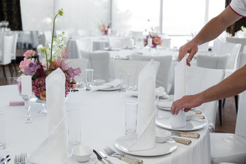 The waiter in the restaurant set up a table for guests. Waiter's hands are a white tablecloth, napkins, white dishes and a bouquet of flowers on the table in the frame