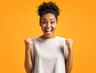 Overjoyed girl clenches fists with happiness, widely opens mouth as shouts loudly. Photo of african american girl wears casual outfit on orange background. Emotions and pleasant feelings concept.