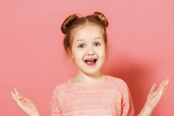 Closeup portrait of attractive charming cheerful little girl with buns over pink background. The child opened his mouth in surprise and spread both hands to the sides.