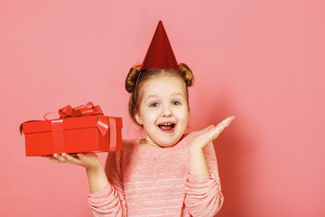 Closeup portrait of a little girl with wisps of hair over pink background. The child has a hat on his head and holds a box with a gift in his hands.