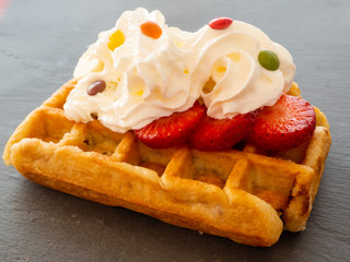 A Belgian waffle with cream and colorful chocolates over strawberry pieces on a slate plate