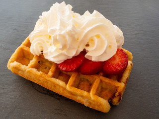 A Belgian waffle with cream over strawberry pieces on a slate plate