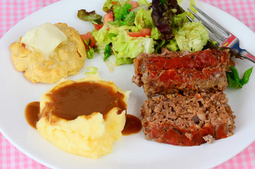 Meatloaf and mashed potatoes on white plate with buttered biscuit and tossed salad.