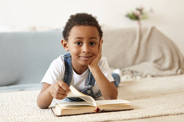 Curious handsome cute African schoolboy reading book on floor in living room. Adorable black male...