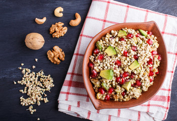 Salad of germinated buckwheat, avocado, walnut and pomegranate seeds in clay plate on black wooden background.
