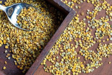 small wooden box containing bee pollen, on a dark wooden table with scattered pollen granules, little spoon inside.