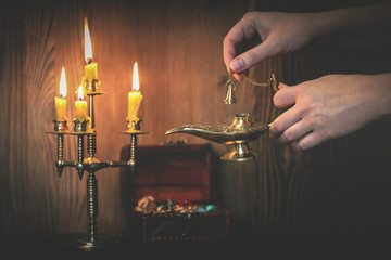 A golden Genie lamp in a woman hands and a treasure chest on a wooden board background in the light of burning candle. Make a wish concept. - Powered by Adobe