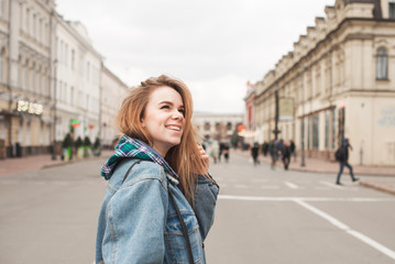 Obraz premium Smiling attractive girl walking along the street of the town, wearing a casual dress jeans jacket, looking sideways, background of the city landscape. Beautiful girl student on the streets of the city