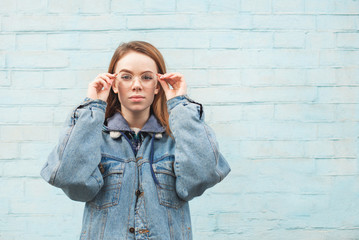 Obraz premium Beautiful girl in a denim jacket stands against the background of a blue wall, looks at the camera and fixes the glasses. Stylish hipster girl posing on a blue background. Copyspace