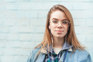 Beautiful girl in the headphones is standing against the background of a blue wall, looking into the camera with a serious face and listening to music. Close up portrait. Copyspace.