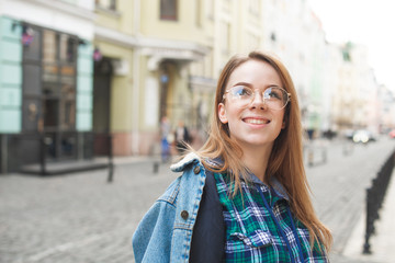 Fototapeta premium Happy student girl in casual clothing, wearing a denim jacket, glasses and a shirt, standing on the back street of the town, looking sideways and smiling. Portrait of a teenage tourist.