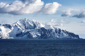 Fototapeta na wymiar Snow peaks, glaciers and rocks of Aleutian islands in sunny winter day as viewed from ship passing in calm sea